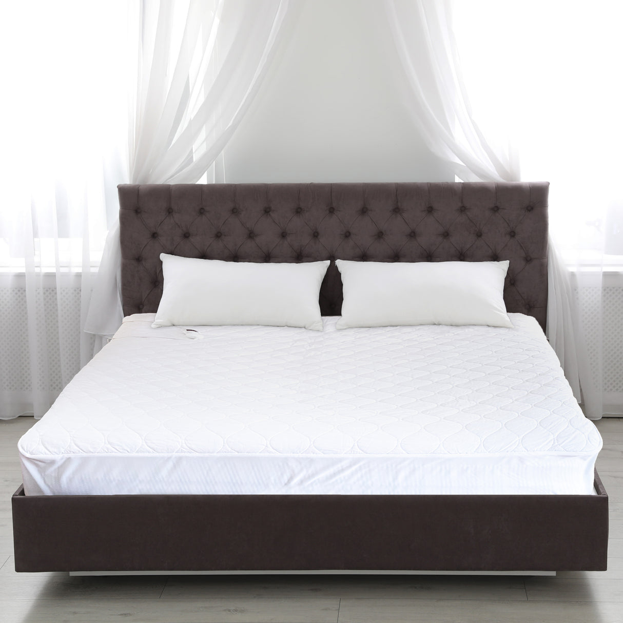 A modern bedroom with a large upholstered bed featuring a dark tufted headboard, a queen-sized mattress topped with the BodyMed Heated Mattress Pad by BodyMed, two white pillows, and sheer white curtains letting in soft natural light.