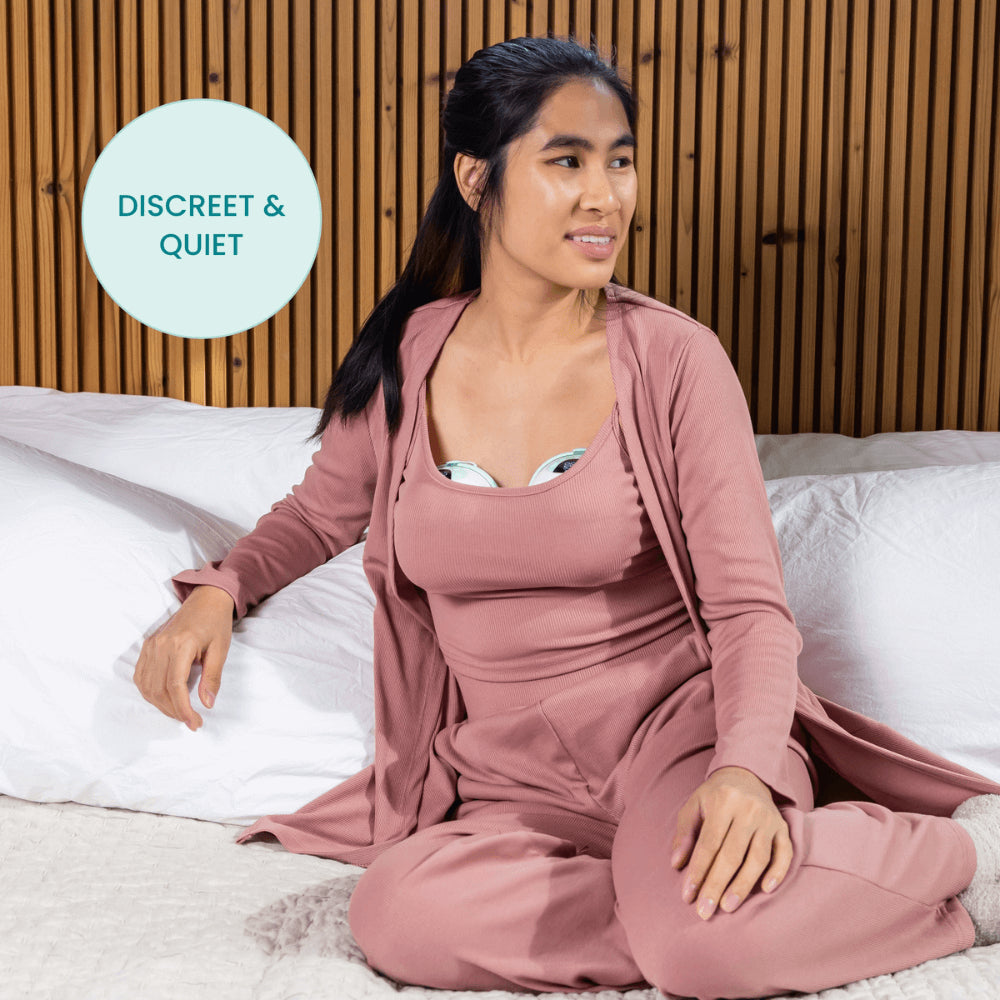 A woman in mauve loungewear sits on a bed, wearing the Motif Aura Glow Wearable Breast Pump by Motif. A circular label reads DISCREET & QUIET. Behind her is a wooden slat wall.