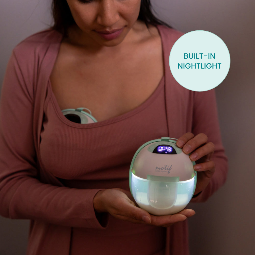 A woman in a pink robe uses the Motif Aura Glow Wearable Breast Pump, featuring a hands-free design, built-in nightlight, and digital display. Another Motif pump is tucked into her bra. A label reads BUILT-IN NIGHTLIGHT.