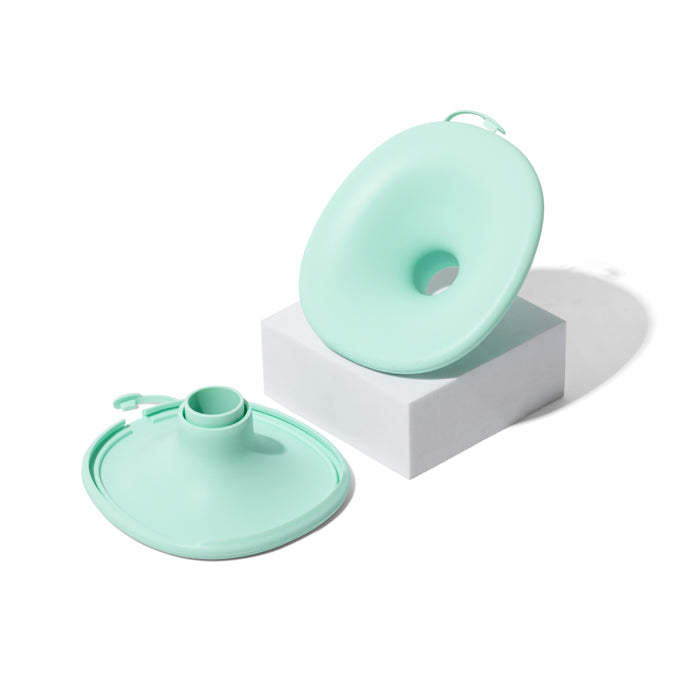 Two mint green, donut-shaped Motif Aura Glow Wearable Breast Pumps by Motif rest on a white block. Each features a small spout and zipper, indicating they are designed to be collapsible or easily refillable.