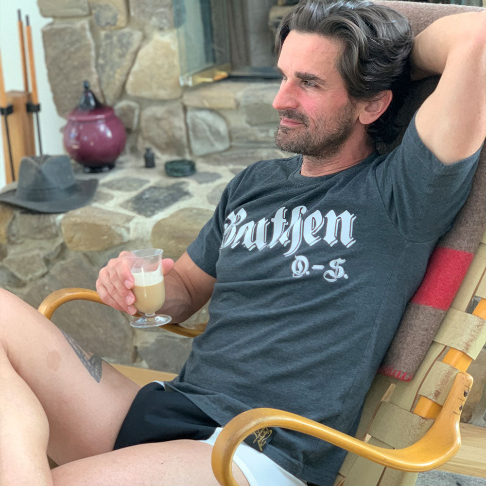 A man with dark hair relaxes in a wooden chair near a stone fireplace, wearing Threaded Armor The Basic Brief, Light Absorbency by ThreadedArmor, enjoying daytime protection. Hes in a dark t-shirt and shorts, holding a drink; a hat and teapot are in the background.