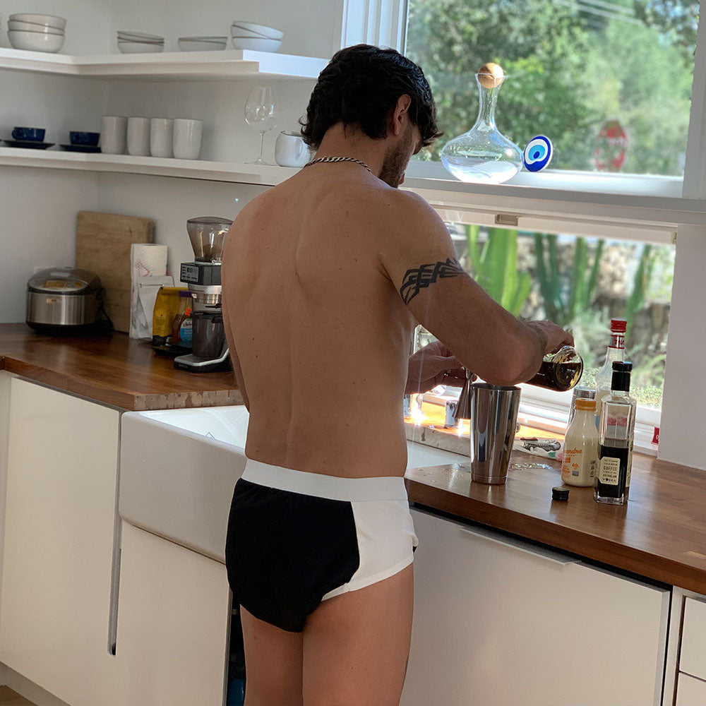 A man wearing ThreadedArmor The Basic Brief, Light Absorbency, stands at a kitchen counter preparing a drink near bottles and a cocktail shaker, sunlight streaming through the window behind him.