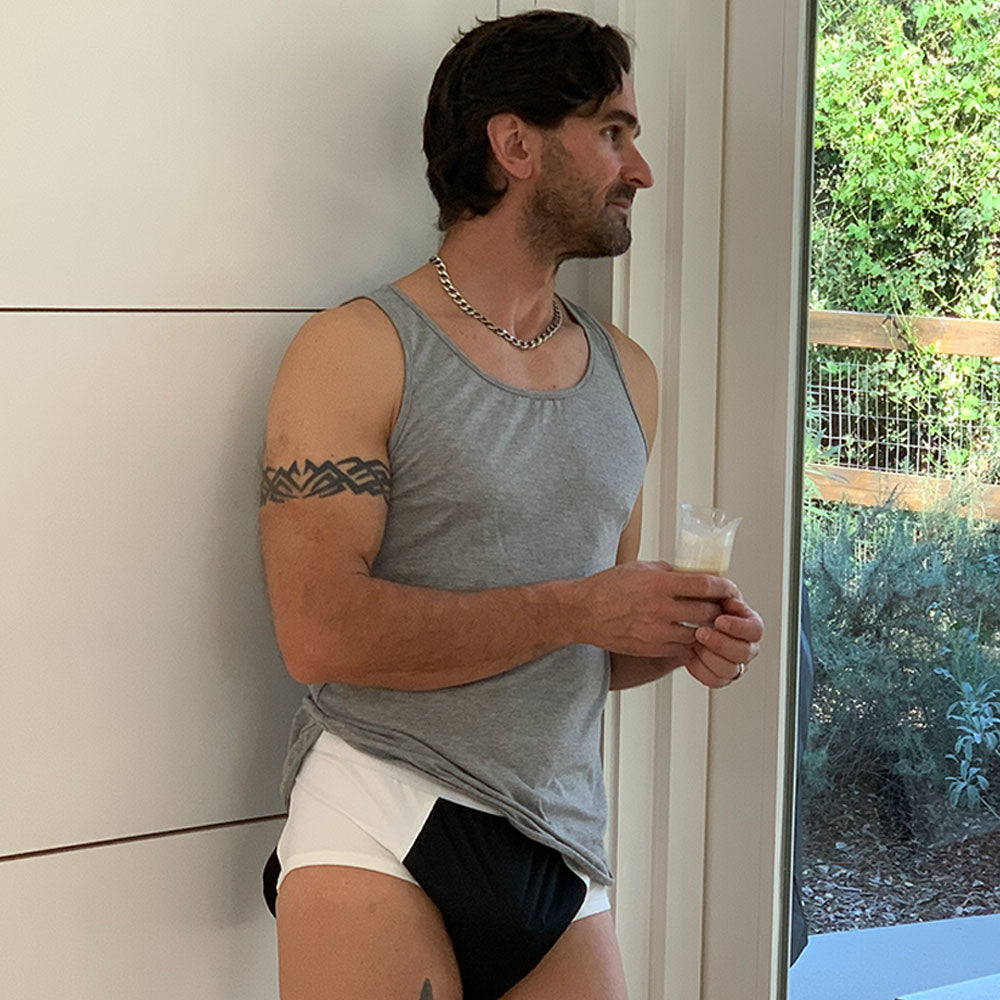 A man in a gray tank top and ThreadedArmor The Basic Brief, Light Absorbency stands indoors by a window, holding a drink and looking outside at green trees and a wooden fence.