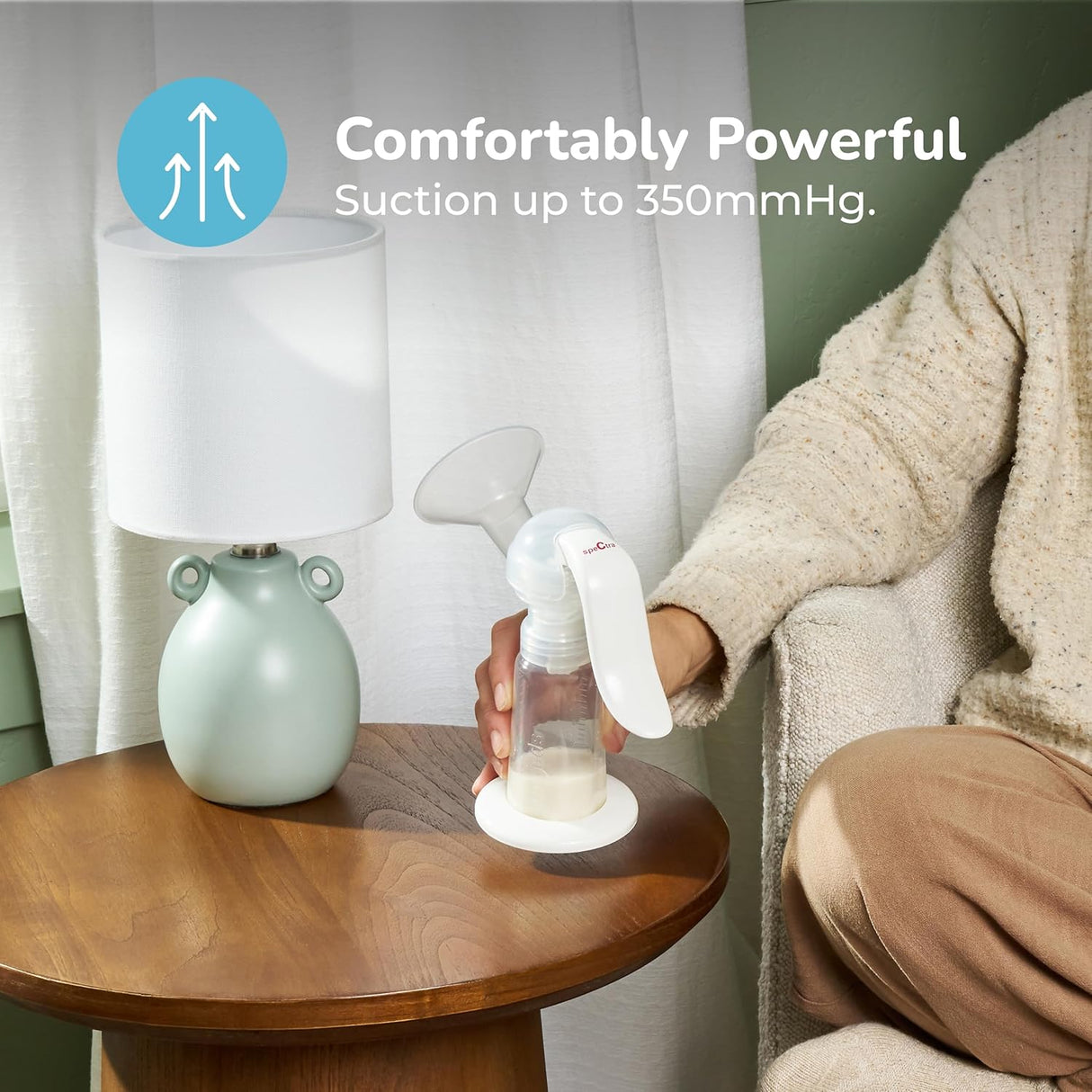 A person uses the Spectra Handy Plus Manual Breast Pump by Spectra while sitting on a beige couch beside a wooden table with a pale green lamp. Text on the image reads, Comfortably Powerful: Suction up to 350mmHg.