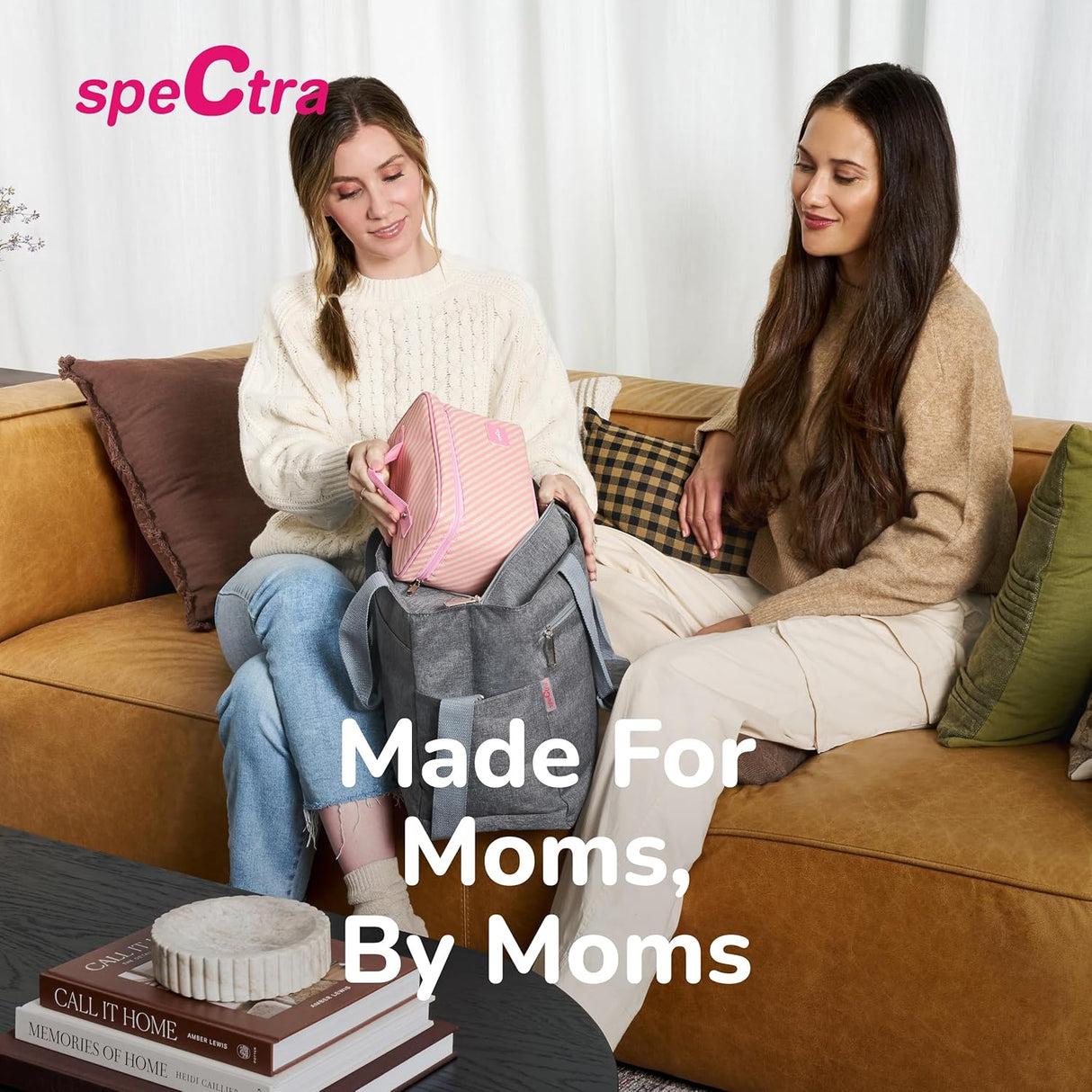 Two women sit on a tan couch. One packs a pink striped pouch into a gray bag, likely her Spectra Handy Plus Manual Breast Pump. The other smiles at her. Text on the image reads Spectra and Made for Moms, By Moms.