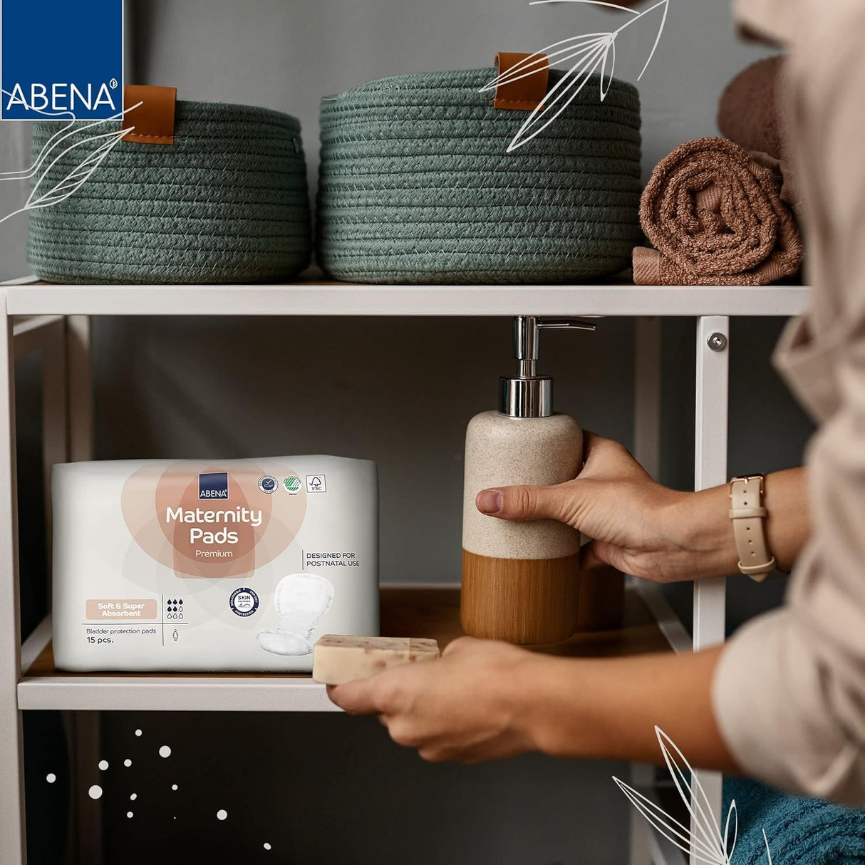 A person sets a soap dispenser on a white shelf beside a pack of Abena Maternity Pads, Super by Abena, made for heavy absorbency, with green and brown woven baskets and rolled towels arranged above.