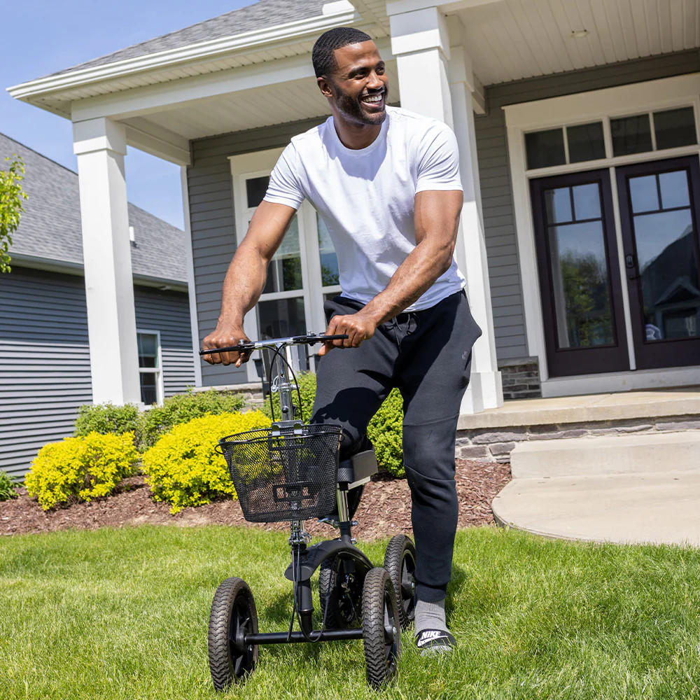 A smiling man in a white T-shirt and black joggers uses the BodyMed All-Terrain Knee Walker by BodyMed on the grass in front of a house with a porch and landscaped yard.