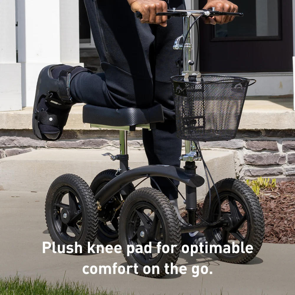 A person with a foot brace rides the BodyMed All-Terrain Knee Walker, featuring a basket and plush knee pad, on a sidewalk in front of a house. Text: Plush knee pad by BodyMed for optimal comfort on the go.