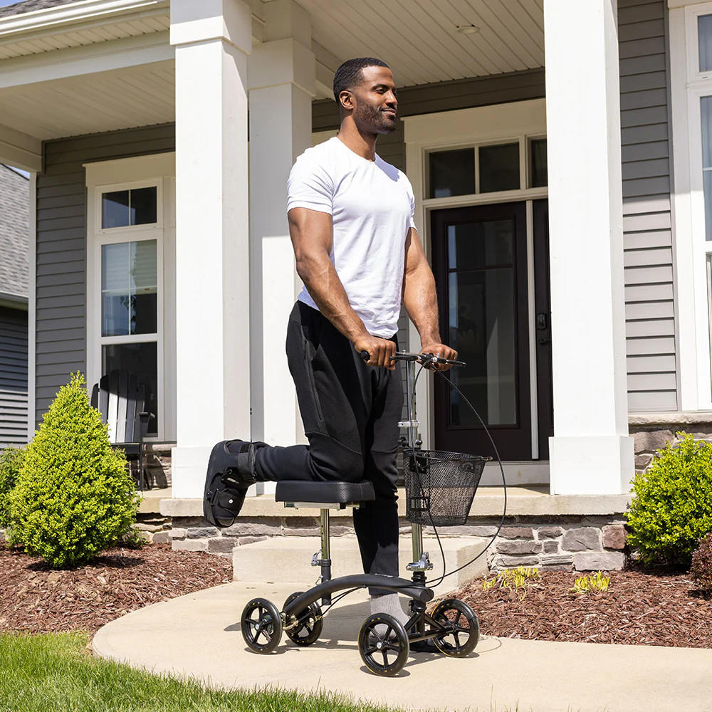 A man uses the BodyMed Premium Knee Walker, a mobility aid by BodyMed, on the sidewalk. He rests his injured right leg on the scooter’s platform and propels himself forward with his left leg while wearing a white T-shirt and black pants.