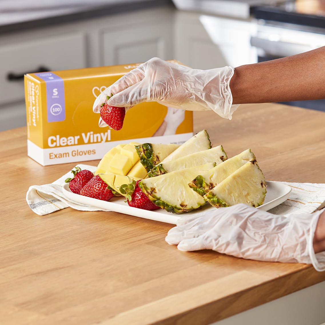 A person wearing CareStock Vinyl Exam Gloves, Standard Cuff, Clear arranges strawberries and pineapple slices on a white plate. A box of CareStock gloves is visible in the background on the kitchen counter.