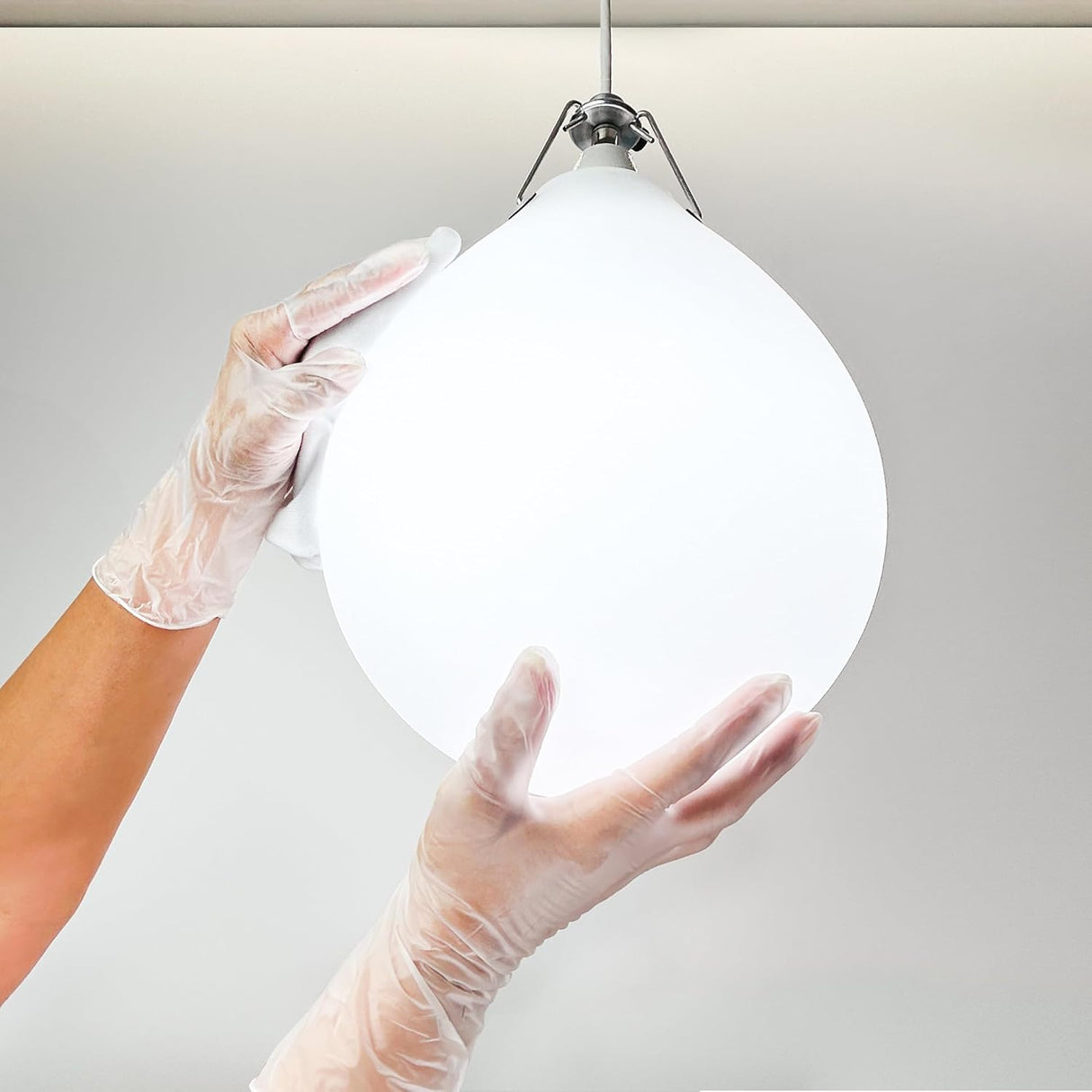 Two hands wearing CareStock Vinyl Exam Gloves, Standard Cuff, Clear by CareStock carefully hold a large, modern, teardrop-shaped white pendant light against a plain, light-colored background.