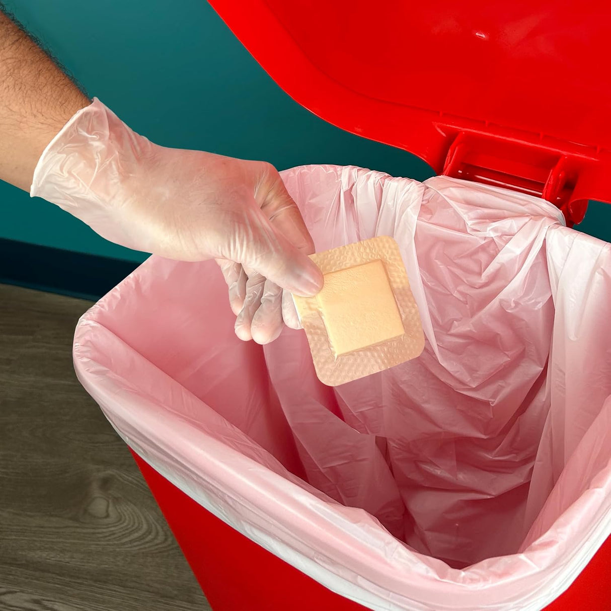 A hand wearing CareStock Vinyl Exam Gloves, Standard Cuff, Clear by CareStock holds a used adhesive bandage above an open red trash bin lined with a pink bag, ready for safe disposal.