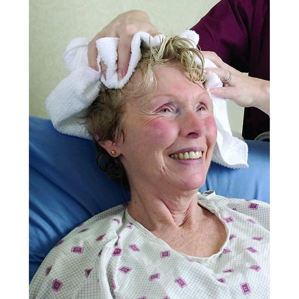 An older woman in a hospital gown smiles as someone gently dries her hair with a white towel and applies CleanLife No Rinse Conditioner, 8 oz, leaving her hair tangle-free. She looks relaxed and comfortable.