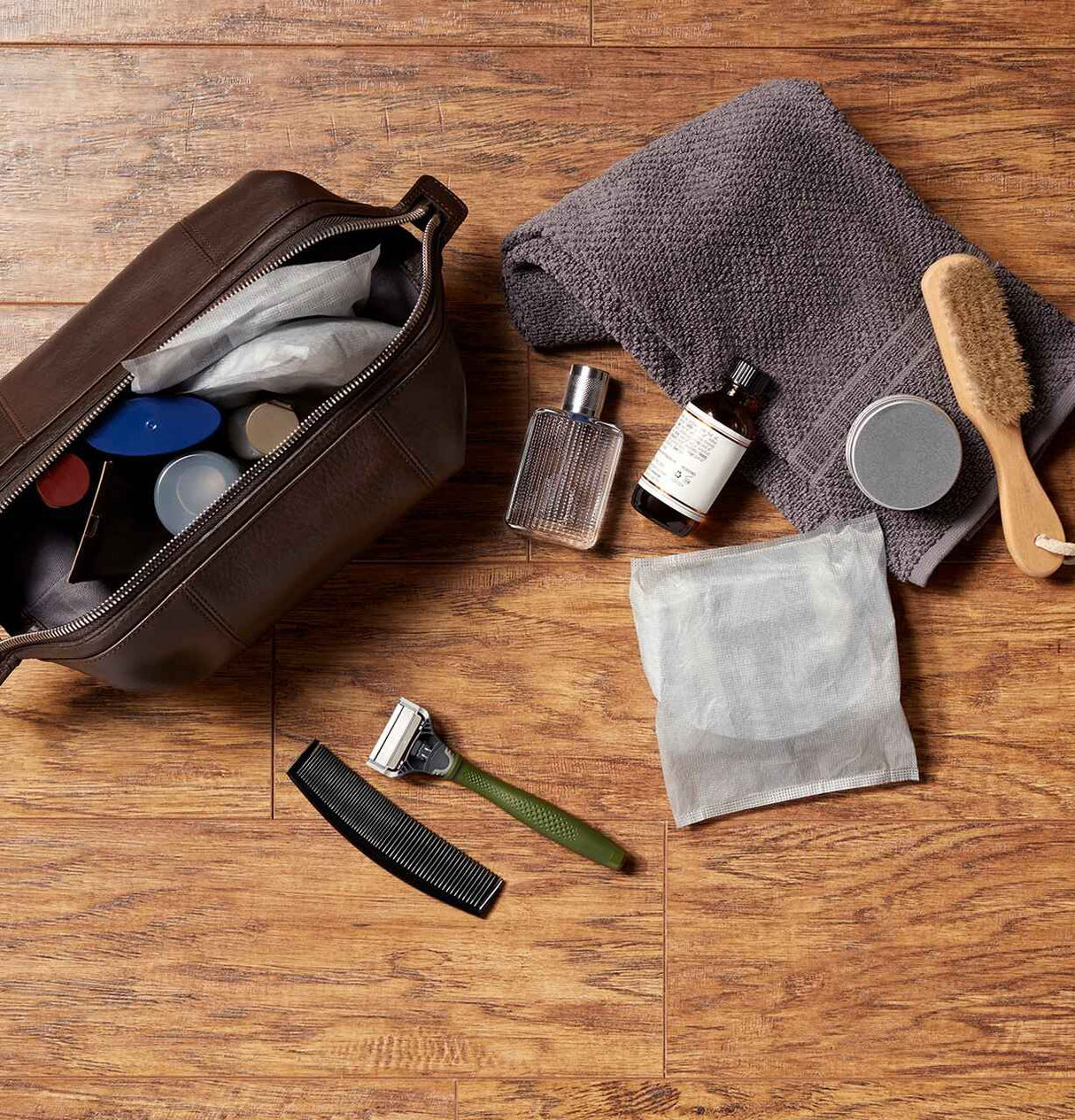 A brown toiletry bag, assorted bottles, a gray towel, a wooden brush, comb, razor, and Depend Incontinence Guards for Men by Depend are arranged on a wooden surface for convenient incontinence protection.