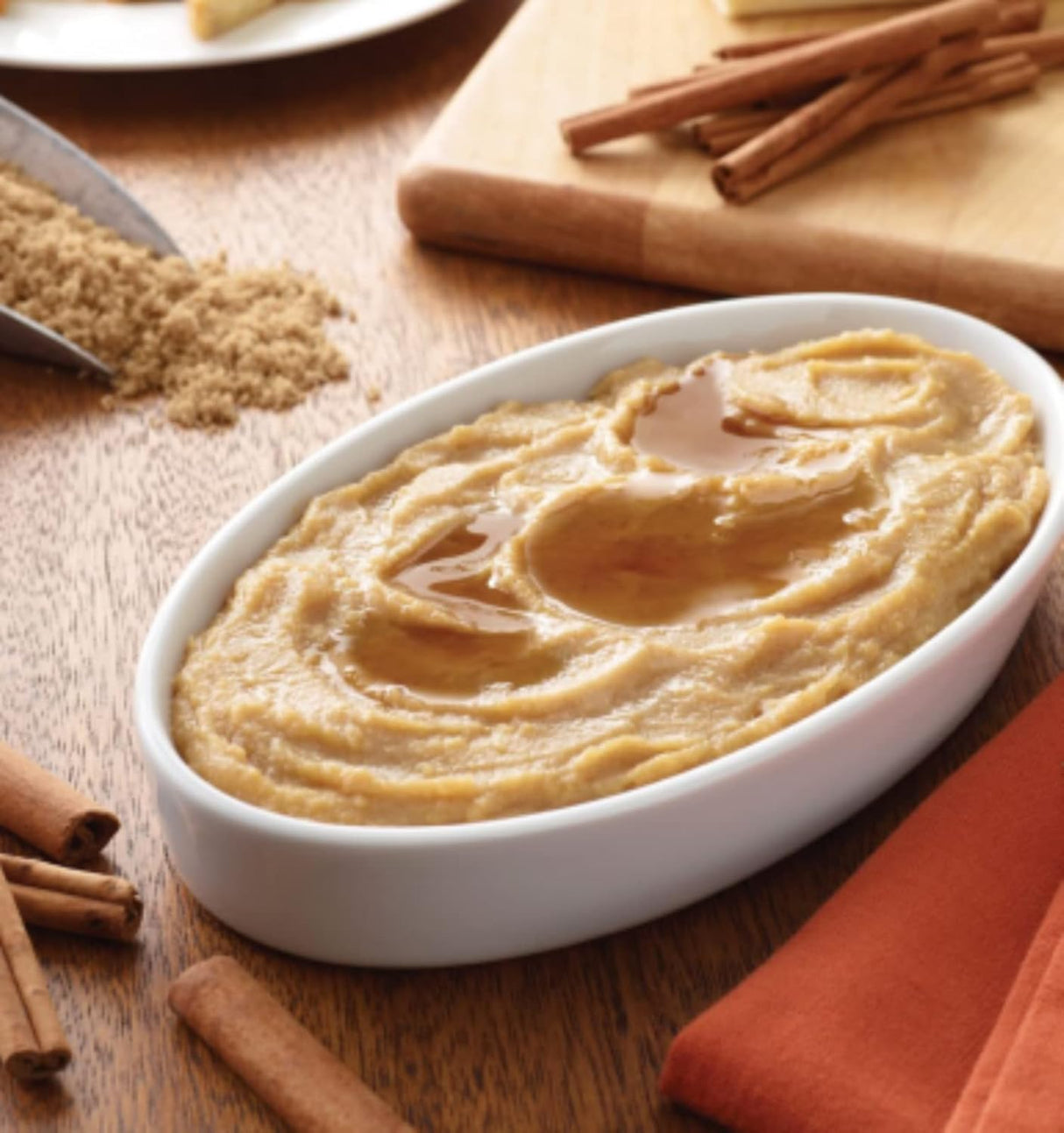 Hormel Thick & Easy Purees Cinnamon French Toast by Hormel Foods is a creamy dysphagia-friendly meal. Served in a white oval dish, its topped with syrup and surrounded by cinnamon sticks, brown sugar, and an orange napkin on a wooden table.