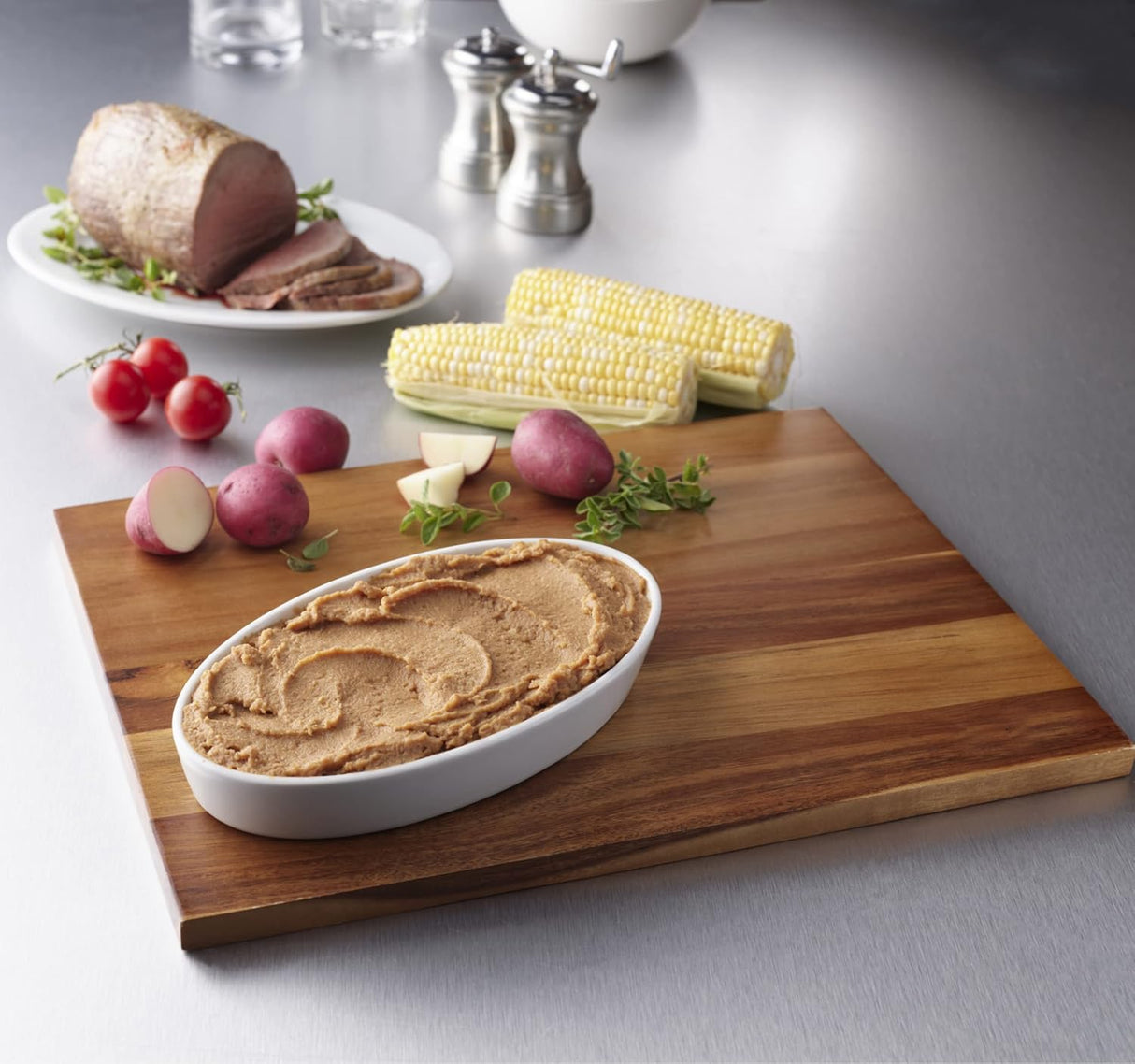 A wooden cutting board with a white dish of whipped butter, red potatoes, radishes, fresh herbs, and corn. In the background is sliced roast beef and Hormel Foods Thick & Easy Purees Homestyle Beef (IDDSI Level 4) on a white plate.