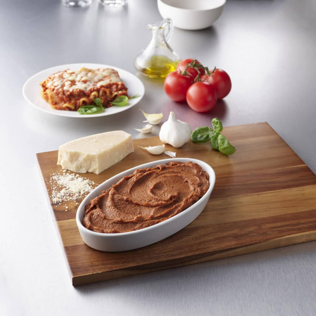 A white dish of tomato paste is on a cutting board with parmesan, garlic, basil, and tomatoes. Nearby, Hormel Thick & Easy Purees Italian Style Lasagna by Hormel Foods (Case/7), a good dysphagia food, sits beside a bottle of olive oil.
