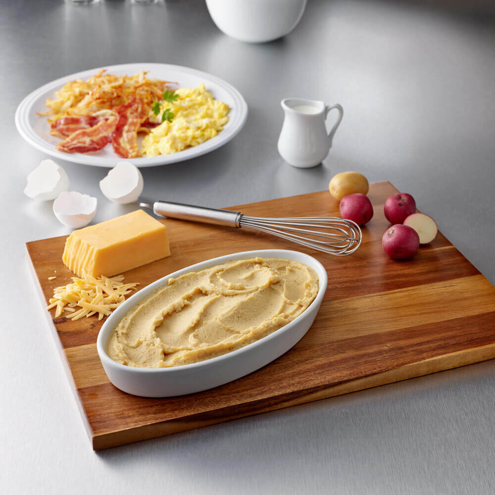 A wooden board holds mashed potatoes, cheddar cheese, a whisk, eggshells, and small potatoes. In the background, Hormel Foods Thick & Easy Purees Scrambled Eggs sit plated with bacon and hash browns next to a small pitcher.