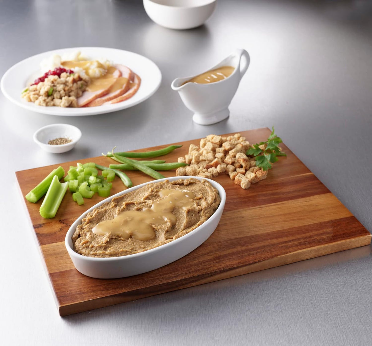 A wooden board displays Hormel Foods Thick & Easy Purees Roasted Turkey topped with gravy, surrounded by celery sticks, green beans, croutons, and parsley. In the background: a plate of IDDSI Level 4 turkey for swallowing difficulties.