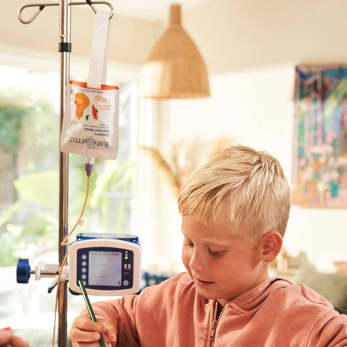 A young blond boy does homework at a table in a cozy, sunlit room while receiving Kate Farms Pediatric Blended Meals, Squash & Carrot, through a feeding tube connected to an IV pole and pump.