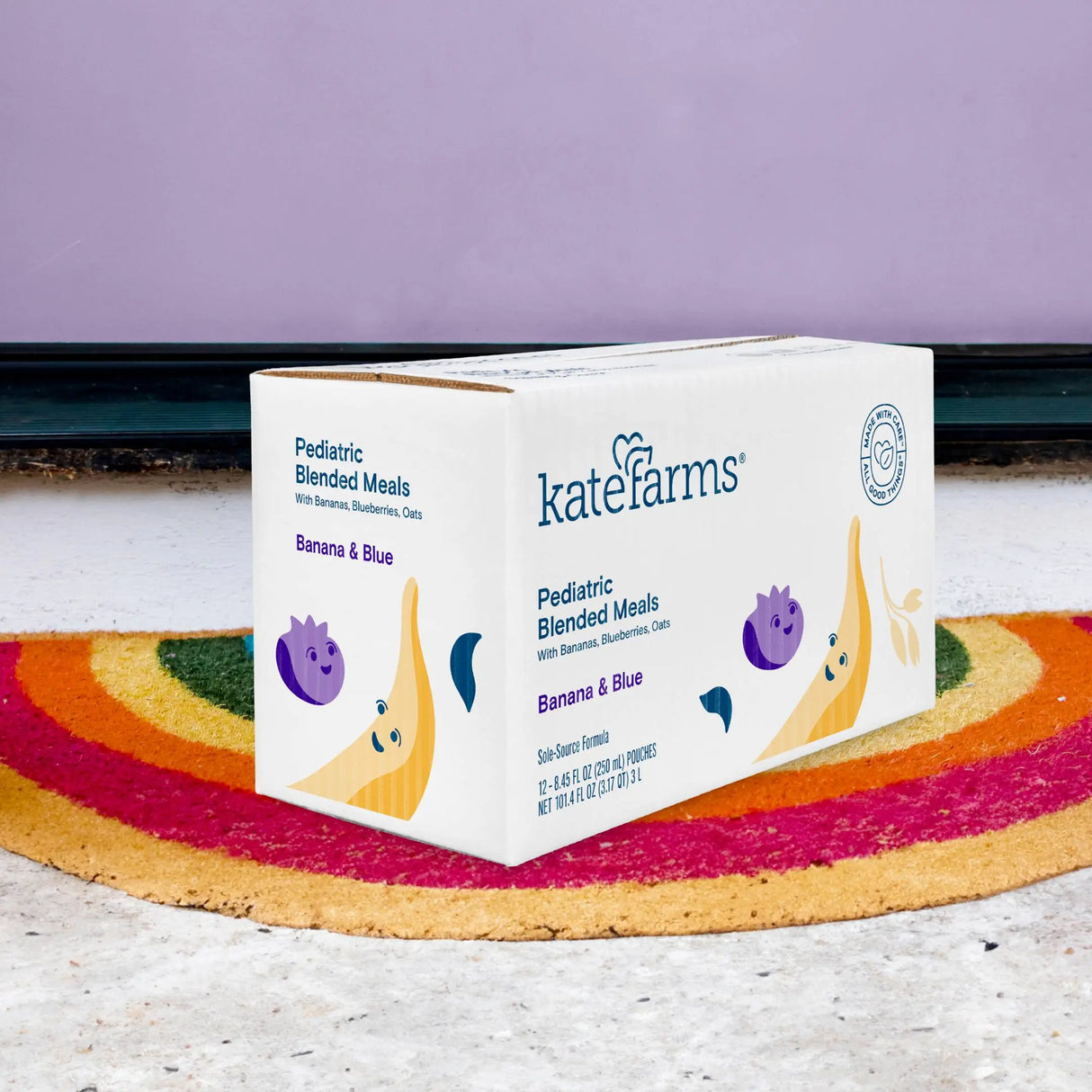 A Kate Farms Pediatric Blended Meals, Banana & Blueberry (CS/12) box sits on a rainbow doormat in front of a purple wall, decorated with banana and blueberry illustrations and containing organic tube feeding formula.