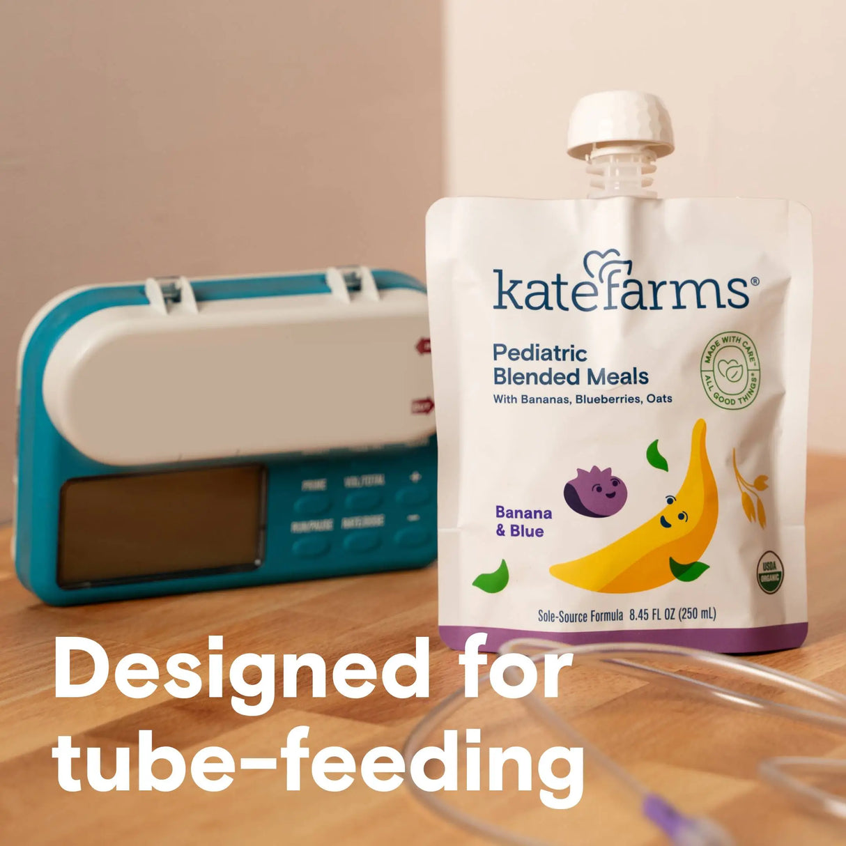 A pouch of Kate Farms Pediatric Blended Meals, Banana & Blueberry, CS/12 sits on a wooden surface next to a medical device, with Designed for tube-feeding text displayed at the bottom.