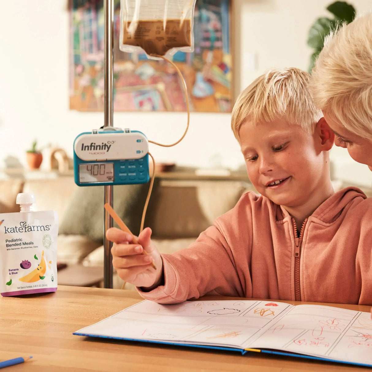 A smiling boy with a feeding tube draws in a notebook at a table, while an adult offers support. Nearby sits a Kate Farms Pediatric Blended Meals, Banana & Blueberry pouch from Kate Farms. The atmosphere is warm and caring.