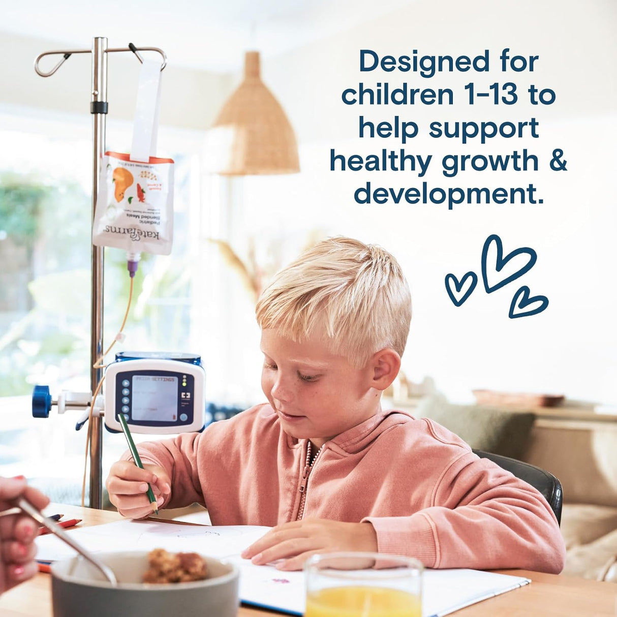 A young child with short blond hair draws at a table beside a Kate Farms Pediatric Blended Meals, Mango & Strawberry pump. Text: Plant-based tube feeding for kids 1-13 supporting healthy growth. Blue heart doodles decorate the scene.