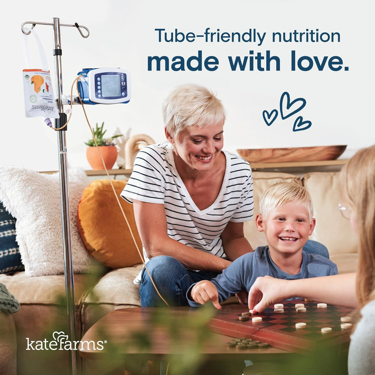 A smiling woman and boy sit on a couch at home with a tube feeding setup. Text reads, Tube-friendly nutrition made with love. Hand-drawn hearts and the Kate Farms logo highlight Kate Farms Pediatric Blended Meals, Mango & Strawberry, CS/12.