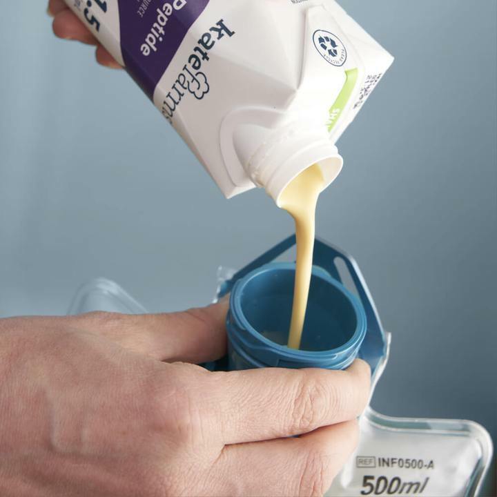 A hand pours thick, beige Kate Farms Peptide Formula 1.5, Vanilla from a carton into a 500ml blue-mouthed medical feeding container, providing complete nutrition.
