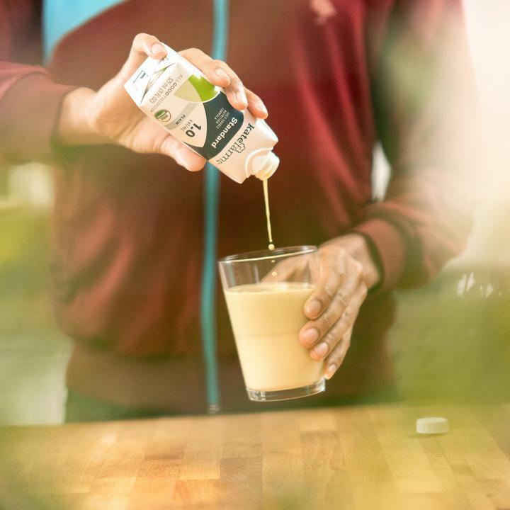 A person in a maroon jacket pours Kate Farms Standard Formula 1.0, Plain from a carton into a glass with a light drink, standing by a wooden table as soft light filters in.