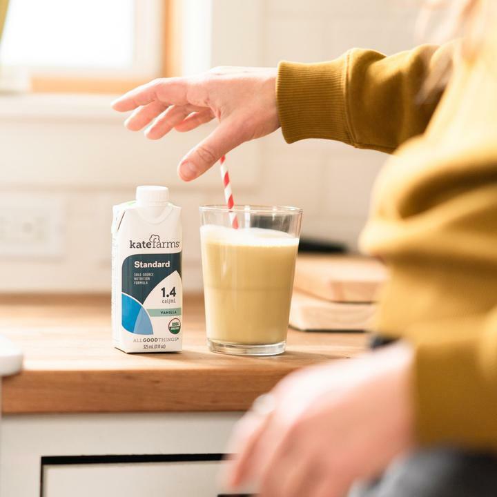 A person in a brown sweater reaches for a creamy vanilla shake with a red-striped straw beside a Kate Farms Standard Formula 1.4, Vanilla carton on the kitchen counter.