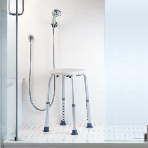 The McKesson Shower Stool, by McKesson, is a white, height-adjustable seat placed in a tiled shower with a handheld showerhead, glass door, and white subway tiles to help promote bath safety.