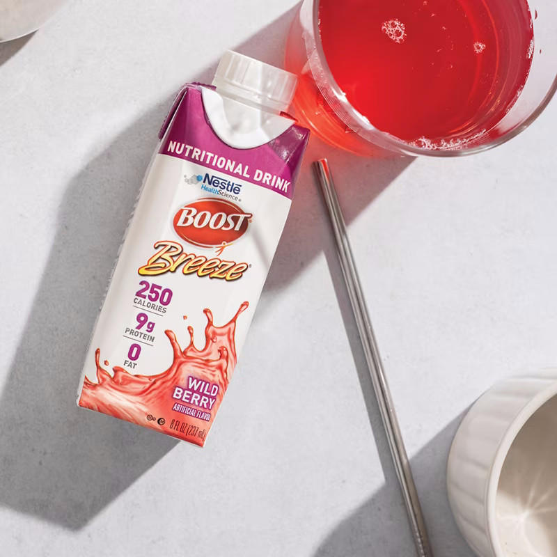 A Nestle Boost Breeze Variety Pack 8 oz carton from Nestle Nutrition is placed next to a metal straw and a glass of bright red drink, all arranged on a light-colored surface.