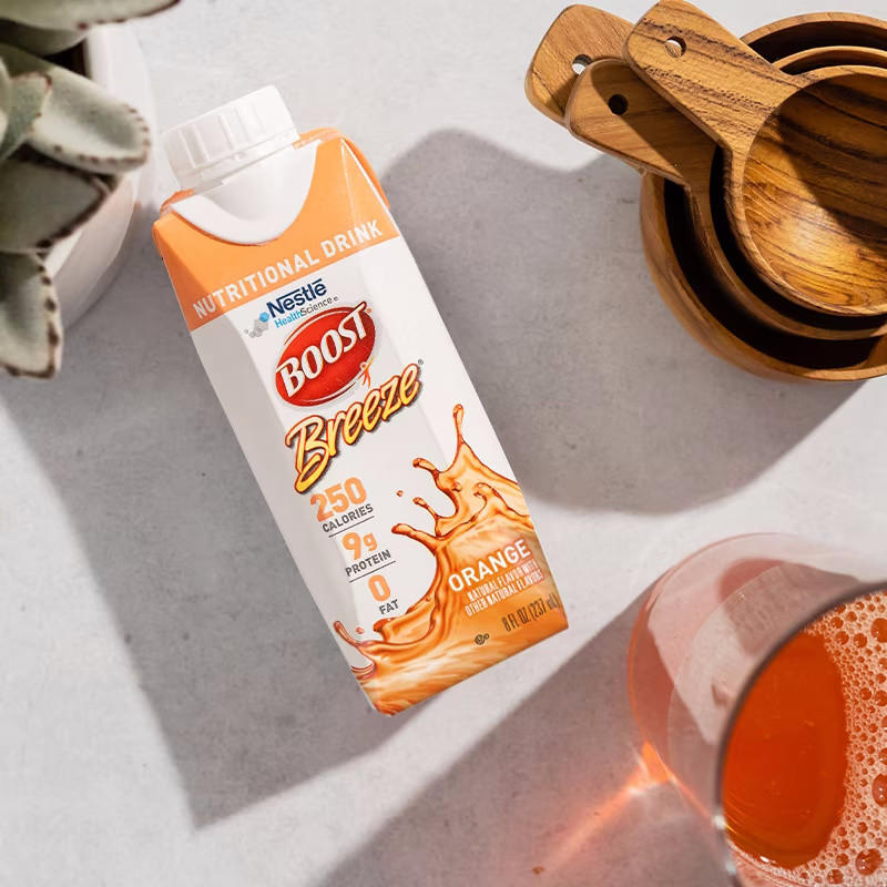 A Nestle Nutrition Boost Breeze Variety Pack 8 oz carton (CS/24) is on a light surface near a glass of orange liquid, a plant, and wooden measuring cups. The carton highlights 250 calories and 9g protein.