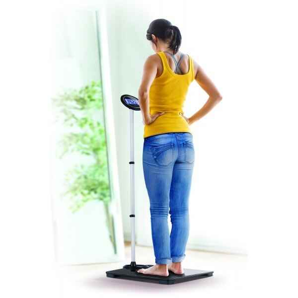 A woman in a yellow tank top and blue jeans stands indoors on the Jobar North American Health + Wellness Extendable Extra Wide Scale, viewing its large LCD display with greenery seen through a bright window in the background.