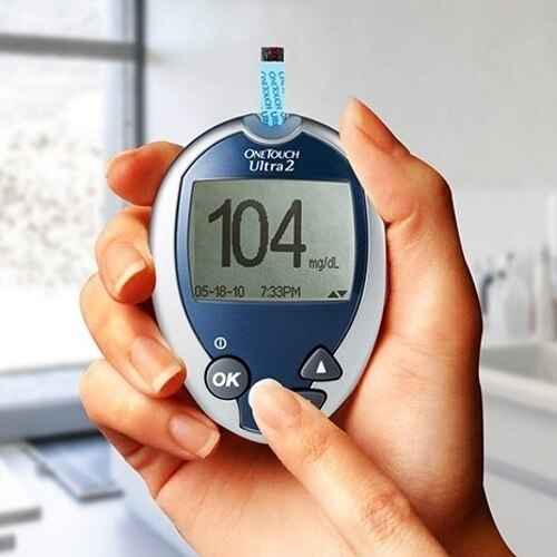 A hand holds a OneTouch Ultra 2 Blood Glucose Meter by OneTouch, showing 104 mg/dL with a test strip inserted, emphasizing diabetes monitoring in a bright kitchen.