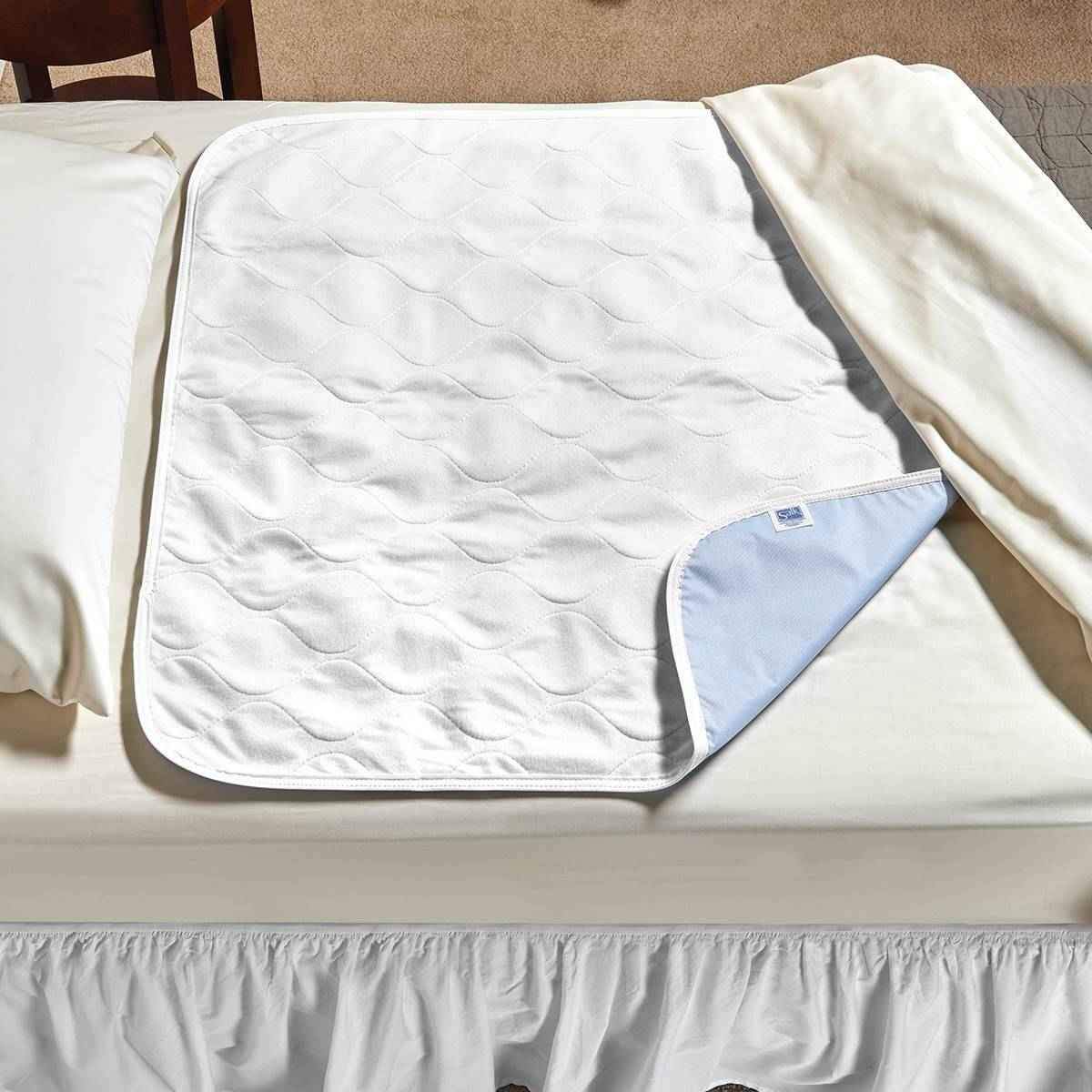 The Salk CareFor Deluxe Underpad by Salk—a white quilted absorbent pad with a light blue backing—lies flat across the center of a neatly made bed, partially tucked under cream-colored sheets.