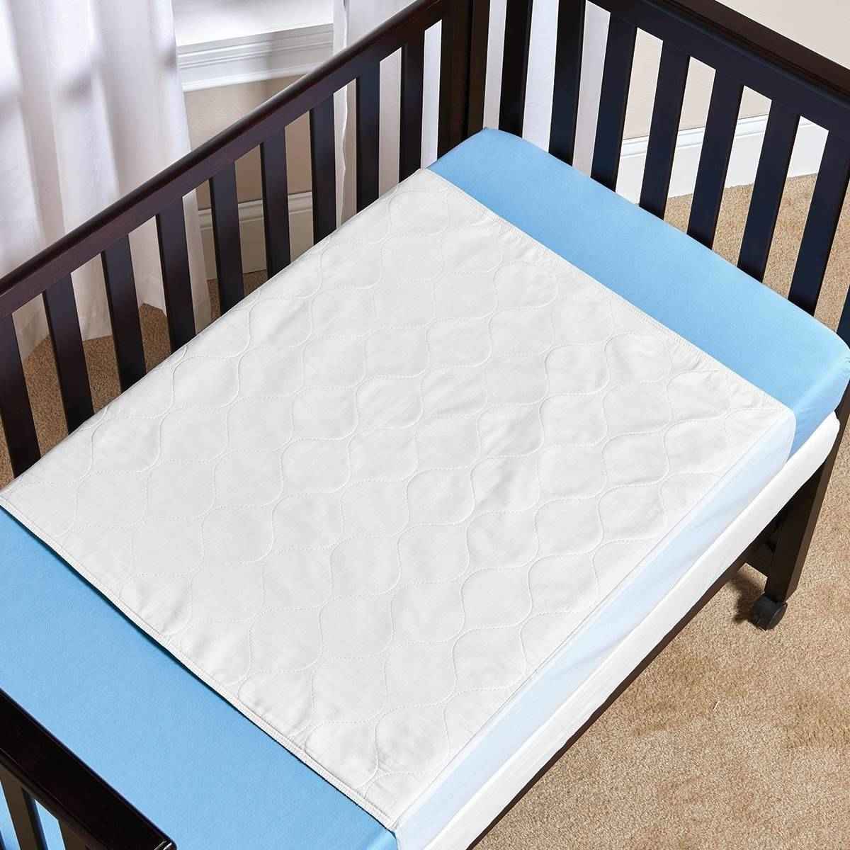 A Salk CareFor Economy Underpad by Salk is placed on a blue crib mattress inside a dark wooden crib, which sits on a beige carpet near white curtains and a wall.