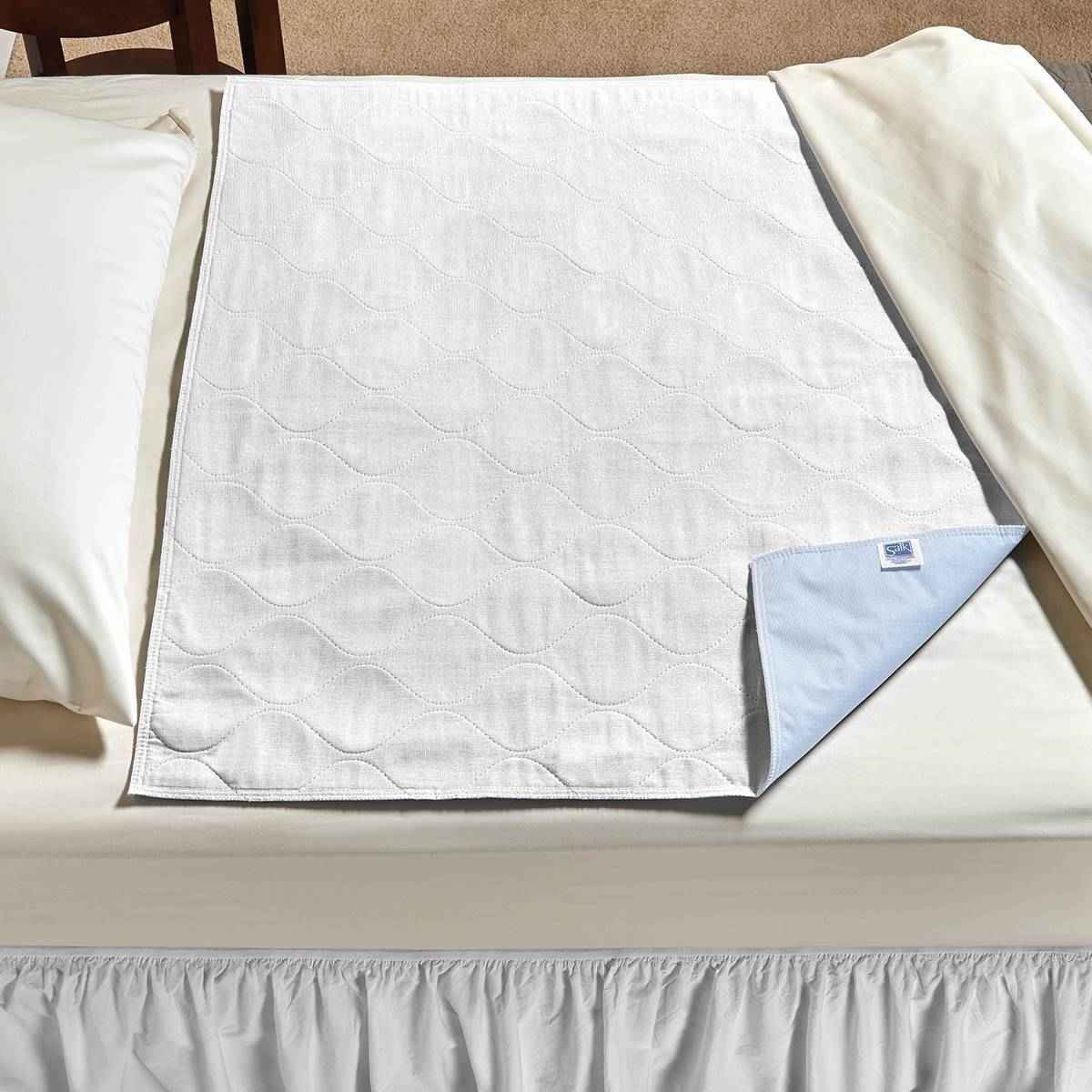 The Salk CareFor Economy Underpad by Salk, a white waterproof pad with a quilted top and blue underside, is partially folded on a made bed between white sheets and a pillow.
