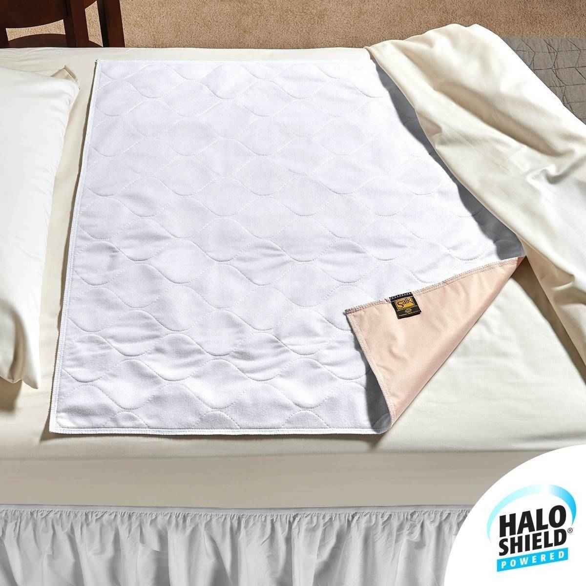A white, rectangular Salk HaloShield Odor Control Underpad with a quilted texture lies on a made bed, one corner folded to show its beige underside. The pad offers antimicrobial protection and features the Halo Shield Powered label in the bottom right.