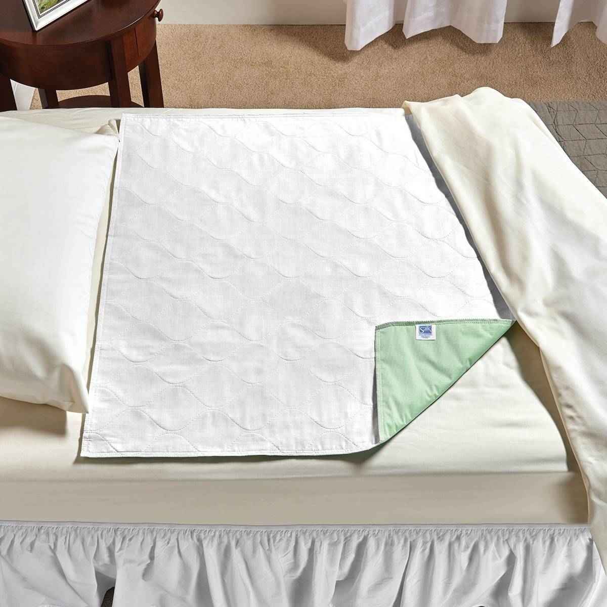 A Salk SleepDri Budget Underpad by Salk, white with a light green underside, is spread over a cream-colored sheet on a bed beside neatly arranged pillows and sheets.