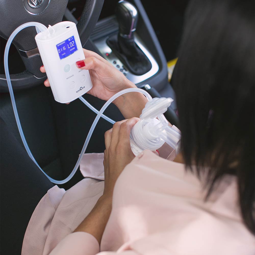 A person in a light pink shirt uses the Spectra 9 Plus Premier Portable Rechargeable Breast Pump by Spectra in their car, holding the compact pump with digital display to their chest.