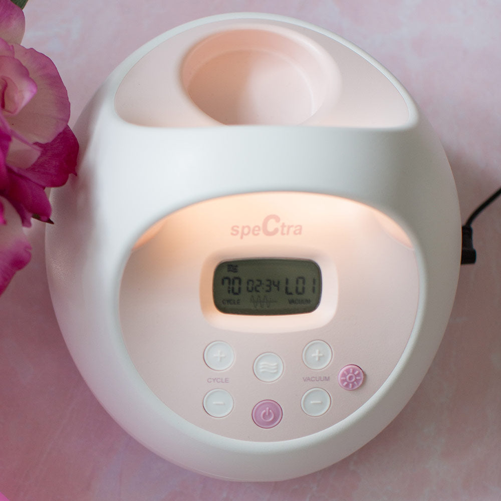 A close-up of the Spectra S2 Plus Double Electric Breast Pump by Spectra, featuring an LCD screen with settings. Pink flowers appear on the left against a light pink background.