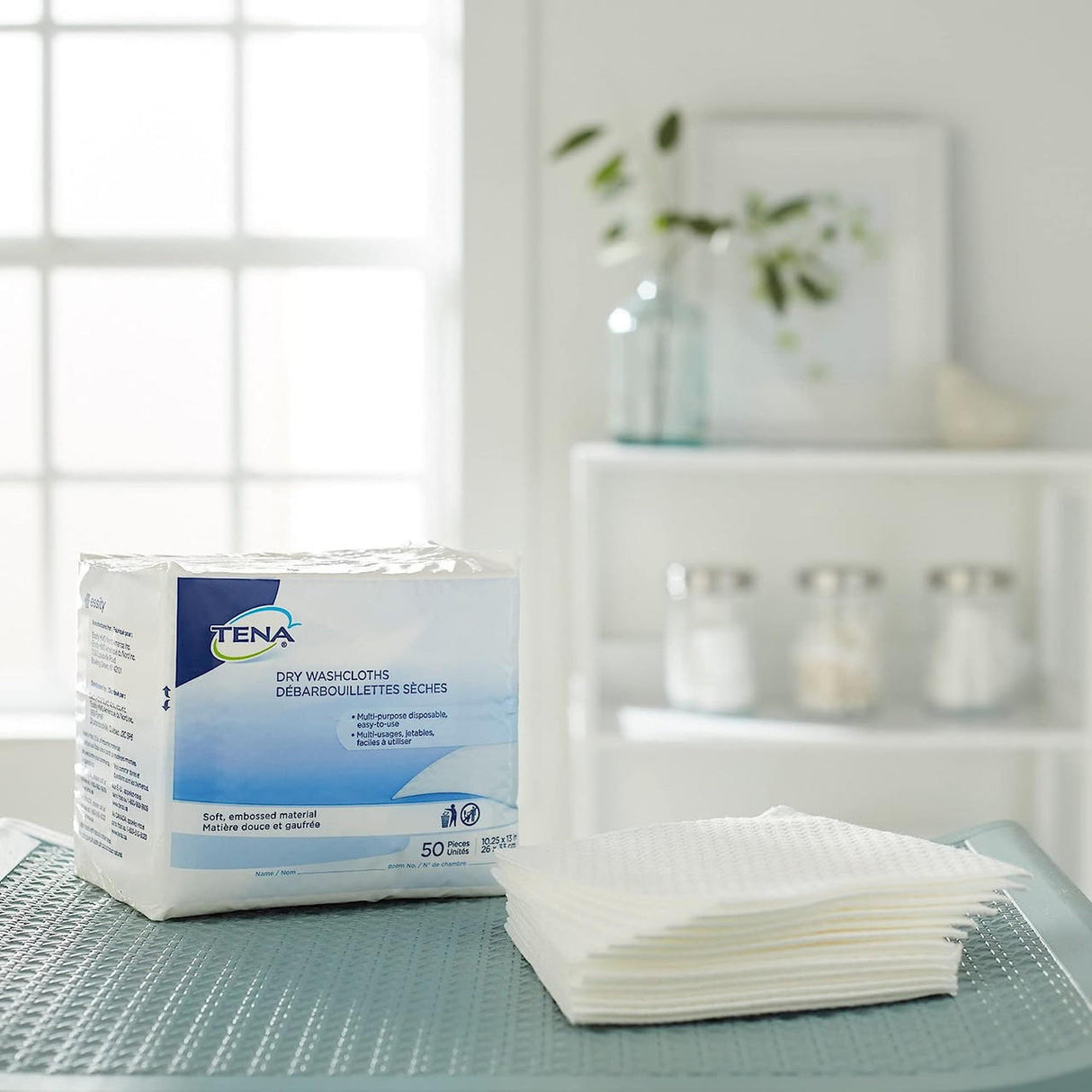 A package of TENA Dry Washcloths by TENA rests on a textured blue mat next to unfolded washcloths in a clean, bright room with shelves, jars, and a plant—ideal for perineal care and everyday hygiene.