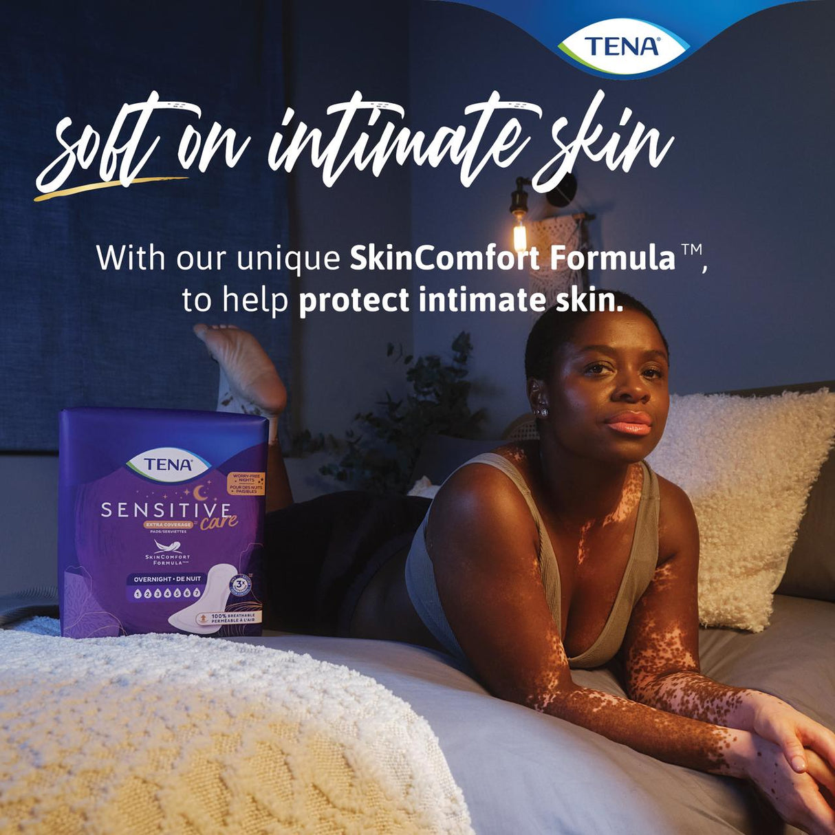A woman with vitiligo lies on a bed beside a box of TENA Sensitive Care Extra Coverage Overnight Pads. Text reads: “Soft on intimate skin. With our unique SkinComfort Formula™, to help protect intimate skin.”.
