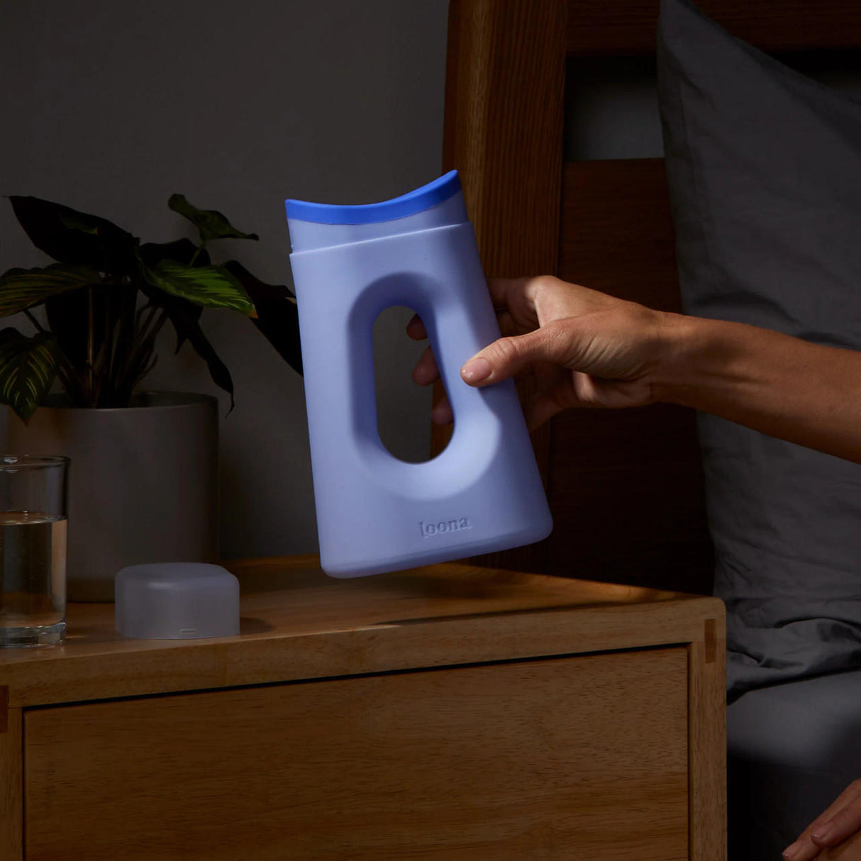 A hand reaches for the Loona Female Urinal With Closure by Boom Home Medical, a blue and white plastic travel urinal with a handle, on a wooden nightstand next to a glass of water, a small container, and a potted plant.
