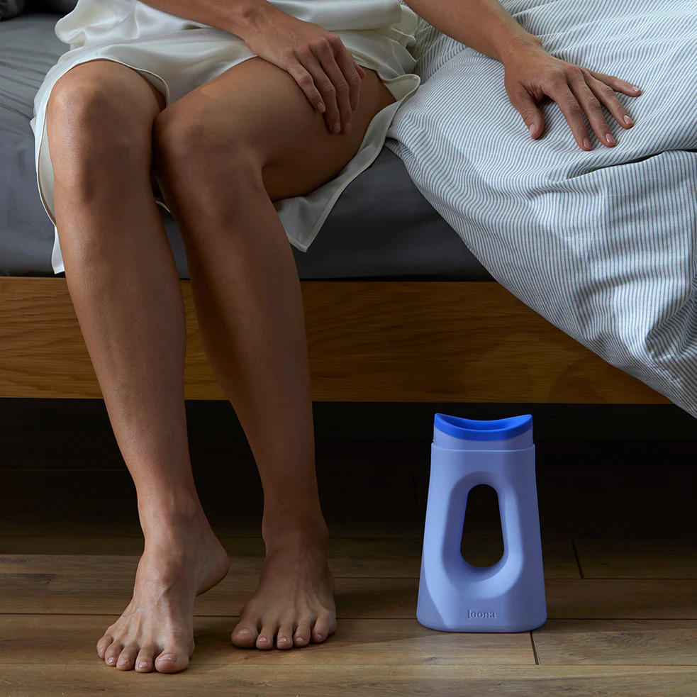 A person in a white nightgown sits on a bed with bare legs visible, and the Boom Home Medical Loona Female Urinal With Closure is placed on the wooden floor beside them.