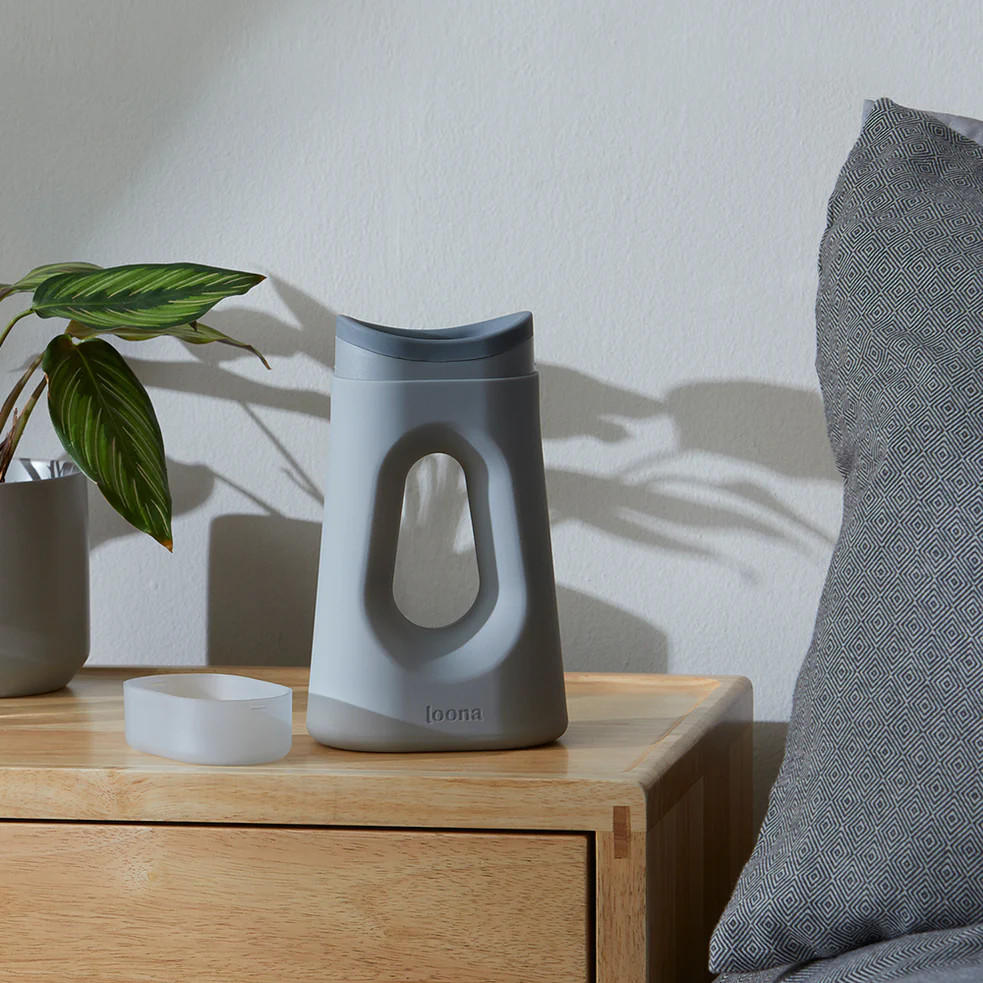 A modern gray tissue dispenser sits on a wooden nightstand beside a potted plant and McKesson Extended Wear Stretch Adult Diapers, with sunlight casting shadows on the wall.