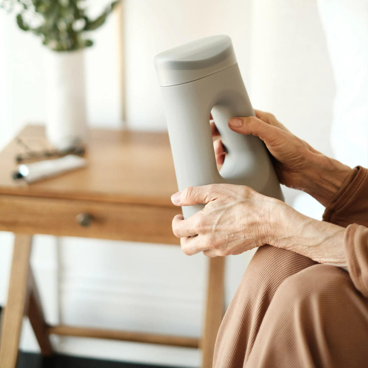 A person in brown holds The Loona Female Urinal With Closure by Boom Home Medical, a modern gray bedside urinal with a handle, sitting next to a wooden side table topped with a plant and tube.